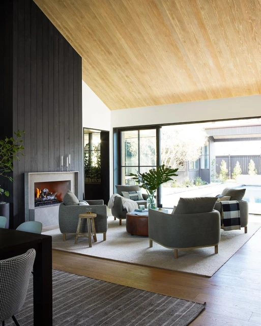 Bright, modern living room with a vaulted wood ceiling, a fireplace, large windows, and gray armchairs arranged around a round ottoman.