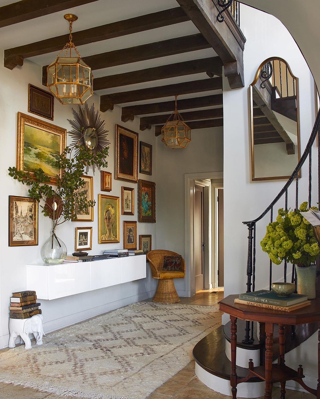 Entryway with vintage-style decor: gallery wall, wicker chair, stacked books on the floor, ornate light fixtures, and patterned rug.