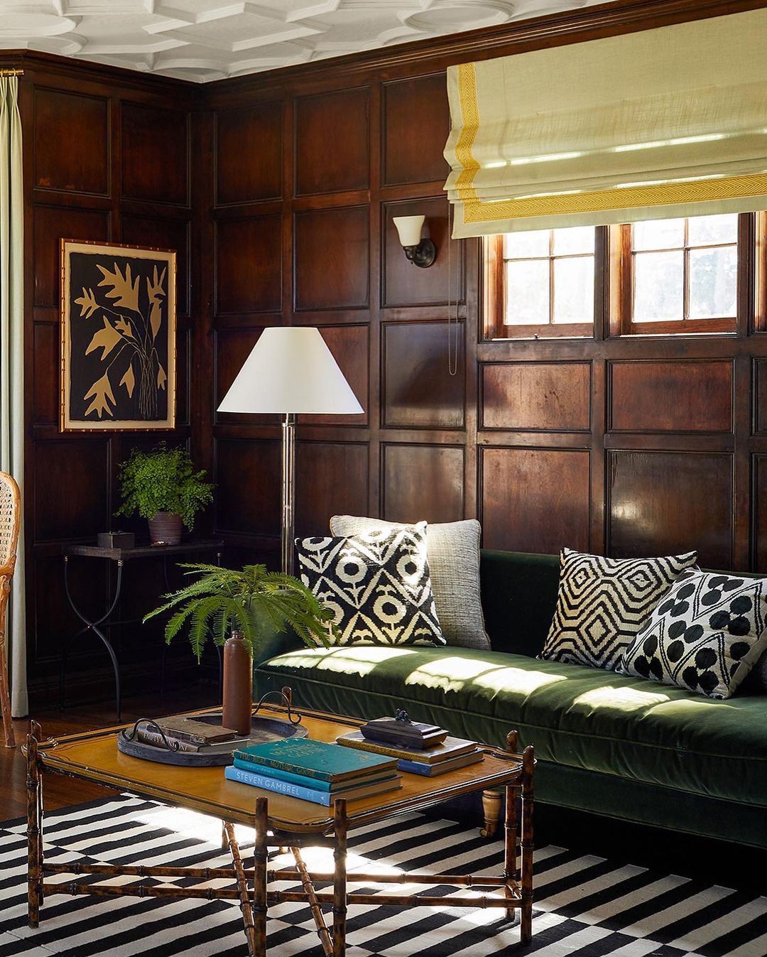 Living room interior with dark wood paneling, green velvet sofa, graphic pillows, patterned rug, coffee table with books and plant.