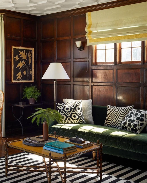 Living room interior with dark wood paneling, green velvet sofa, graphic pillows, patterned rug, coffee table with books and plant.