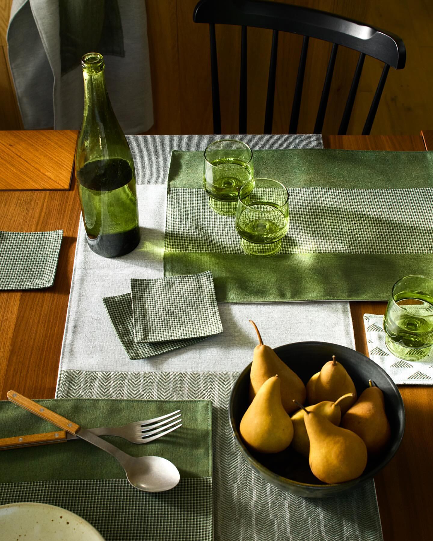 Table setting with pears, green glassware, and linens in natural light, arranged on a wooden table with a black chair in background.