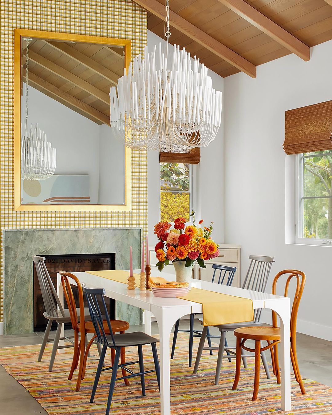 Dining room interior features a white table, assorted chairs, modern chandelier, floral centerpiece, and gold framed mirror on a patterned wall.