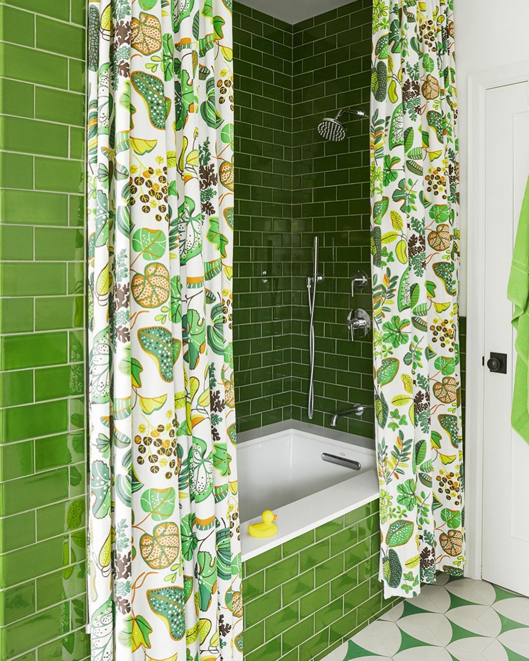 Bathroom interior with green tile walls, floral shower curtain, white bathtub, and round-patterned floor tile. Bathtub has a yellow duck.