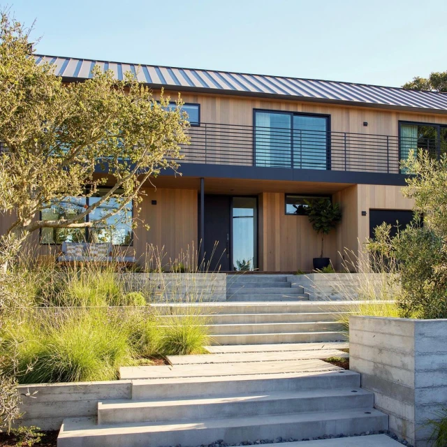 Exterior of a modern two-story house with light wood siding, a dark metal roof, a second-story balcony, and a concrete stairway.