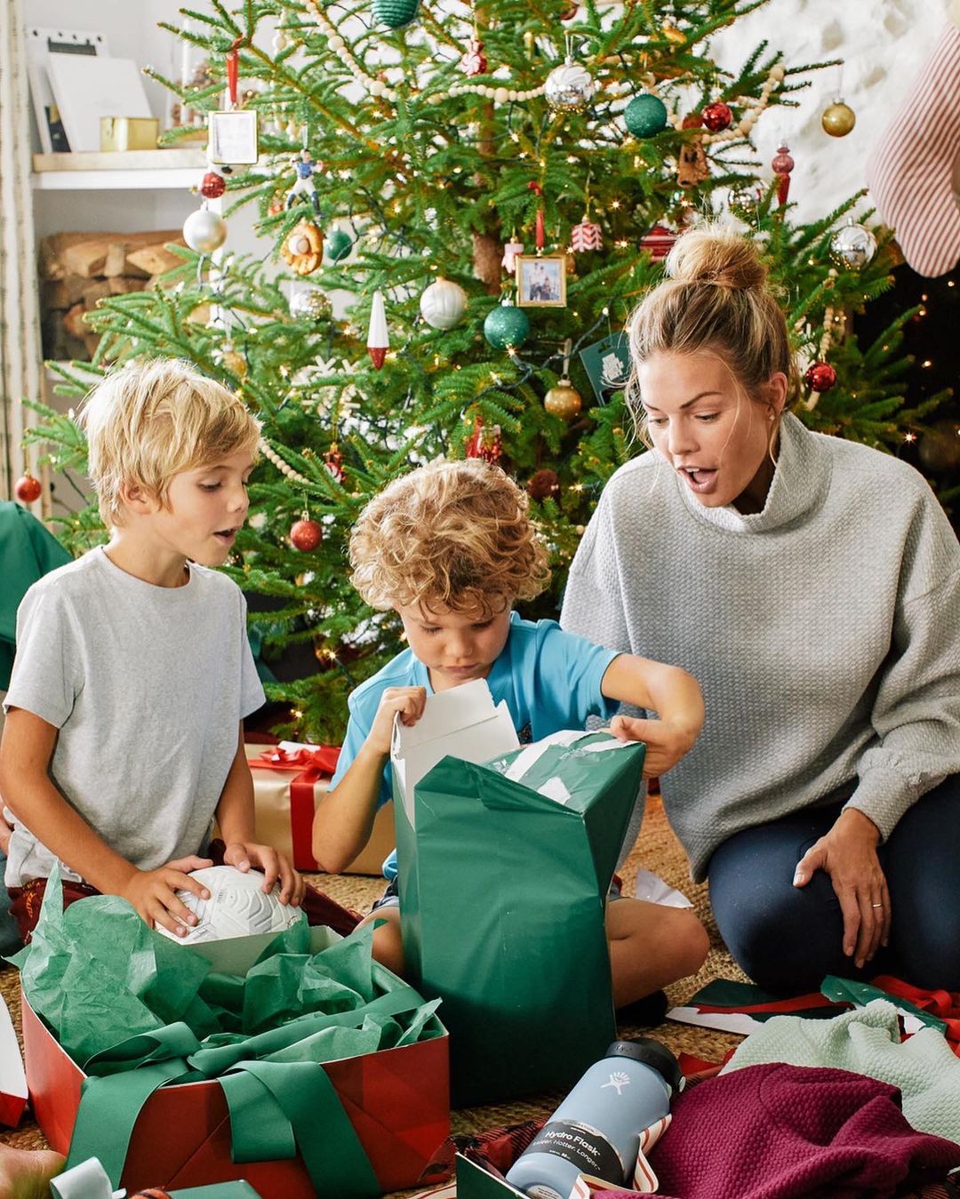 Mother and two children unwrapping gifts near a Christmas tree in a cozy home interior with presents and holiday decorations.