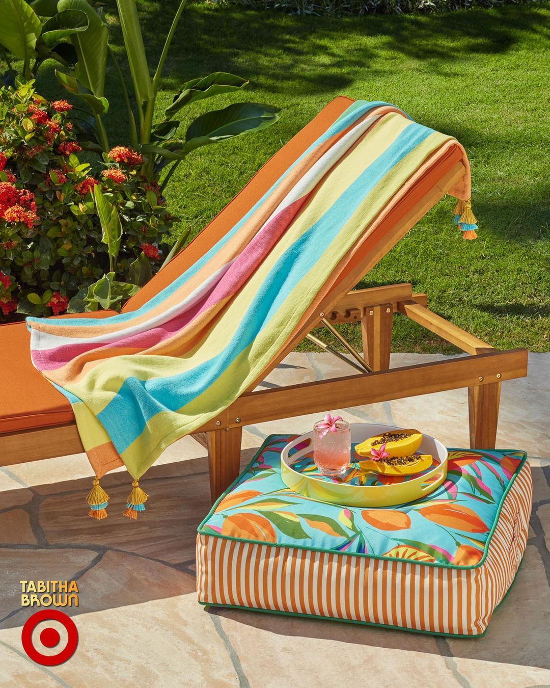 Outdoor setup: wooden chaise lounge with a colorful striped towel, a patterned ottoman with a tray holding drinks and sliced fruit.