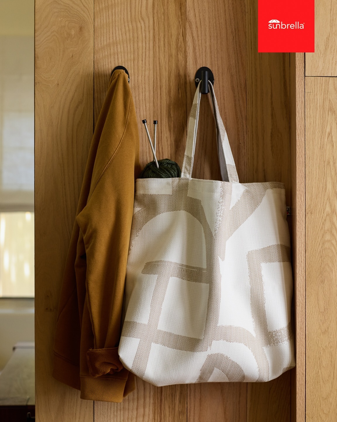 Tote bag with a geometric pattern hanging on a wooden wall, alongside a mustard-colored jacket and knitting yarn, in an indoor setting.