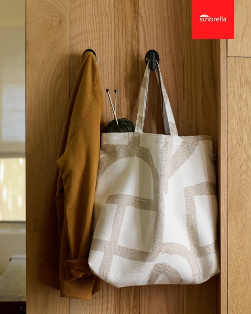 Tote bag with a geometric pattern hanging on a wooden wall, alongside a mustard-colored jacket and knitting yarn, in an indoor setting.