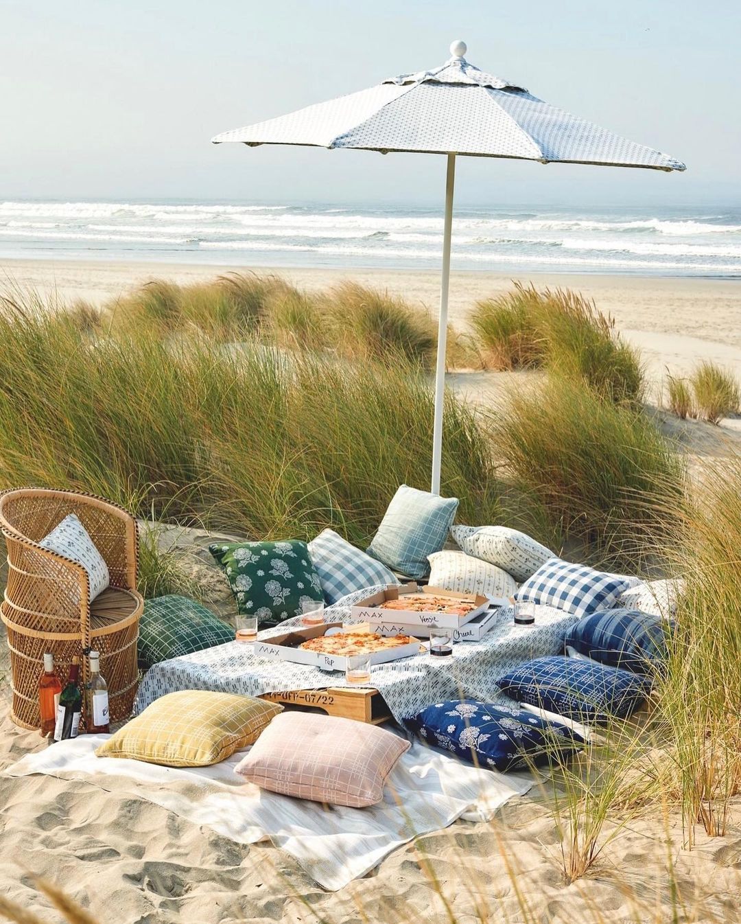 Picnic setup on a sandy beach with a table covered with pizza boxes and pillows, sheltered by a white parasol, with the ocean in the background.