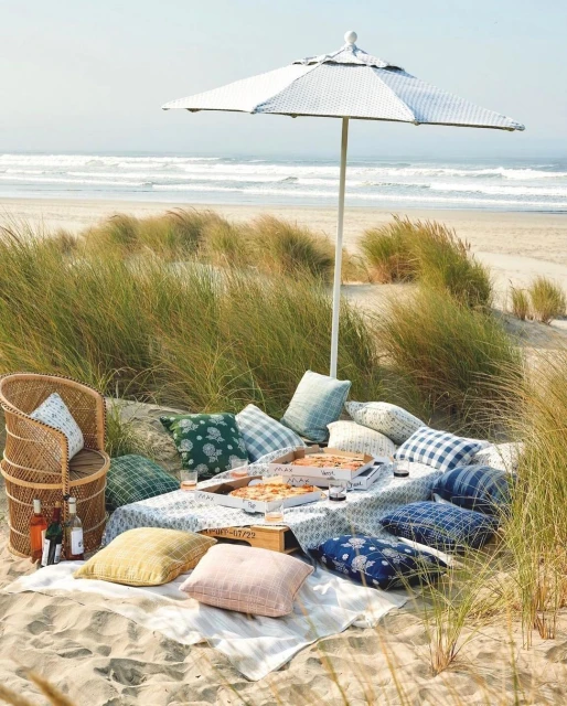 Picnic setup on a sandy beach with a table covered with pizza boxes and pillows, sheltered by a white parasol, with the ocean in the background.