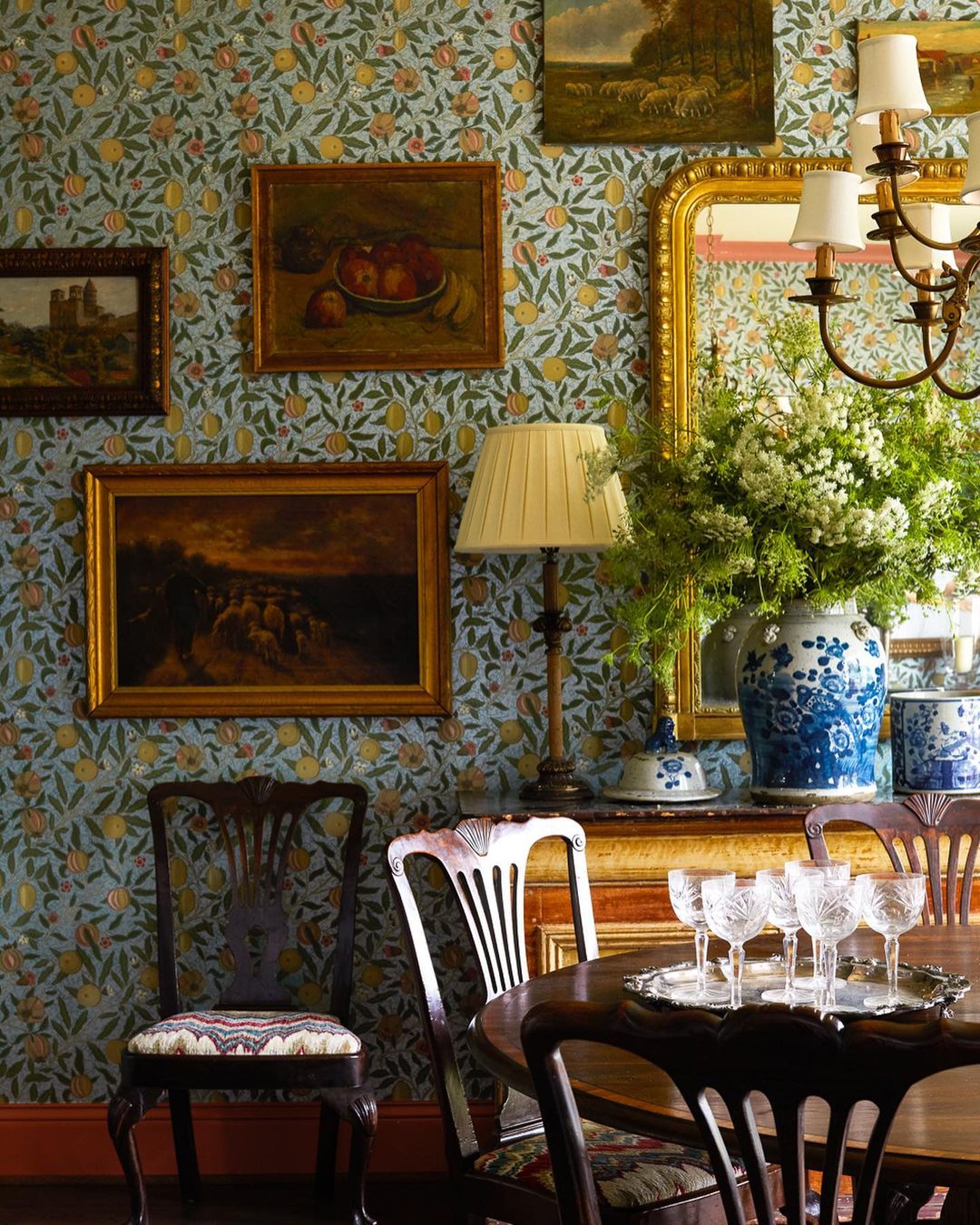 Interior view of a dining room with floral wallpaper, antique furniture, artwork, a table setting with glassware, and an arrangement.