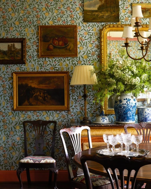 Interior view of a dining room with floral wallpaper, antique furniture, artwork, a table setting with glassware, and an arrangement.