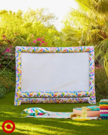 Outdoor inflatable movie screen on green lawn surrounded by trees. A picnic blanket and seating are arranged in front of the screen.