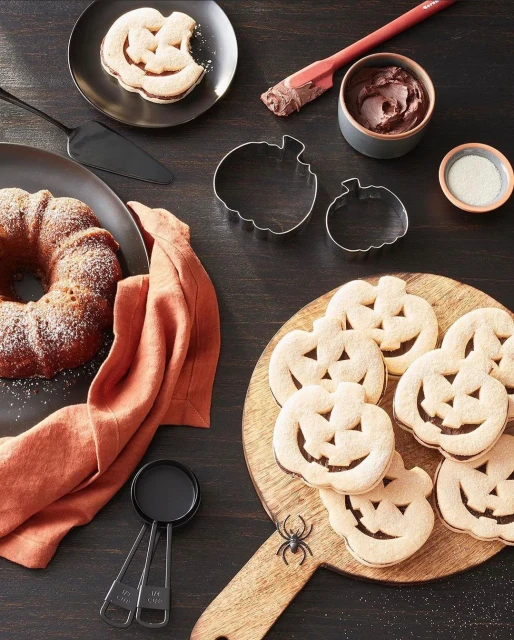 Overhead shot of Halloween-themed desserts including pumpkin cookies, a bundt cake, and ingredients, set on a dark wooden surface.