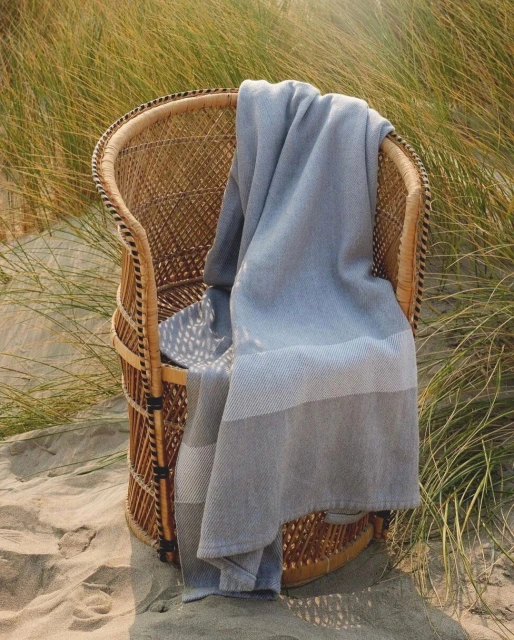 Wicker chair on sandy beach with blue and gray striped towel draped over it, with natural beach vegetation in the background.