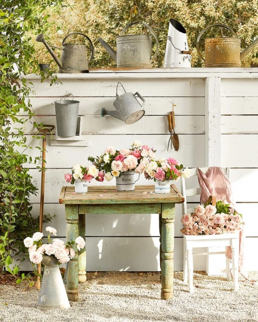 Collection of vintage watering cans on a shelf above a rustic green table with flowers in a garden setting with white walls.