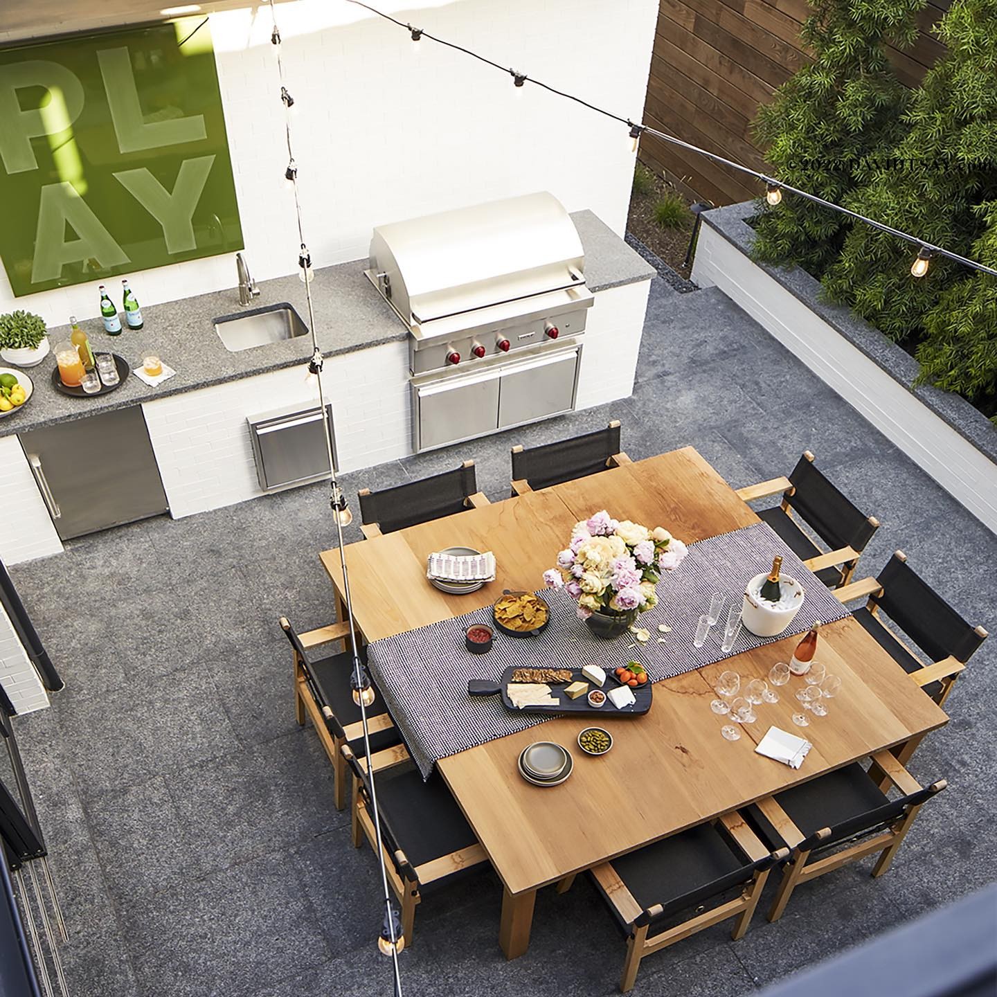 Outdoor patio with a wooden dining table set for a meal, an outdoor kitchen, grill, string lights, and leafy green plants.