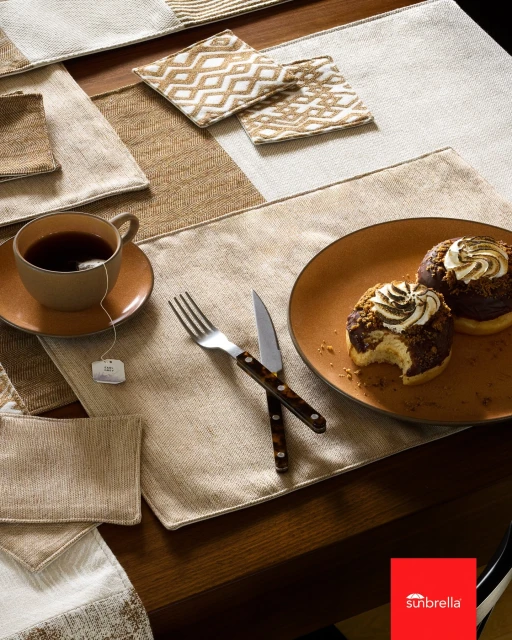 Table setting featuring neutral-toned placemats, a cup of tea, an orange plate with two donuts, and silverware on a dark wood table.
