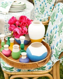 Colorful ceramic vases and candleholders on a woven rattan tray resting on a chair with blue and green floral cushions, photographed by David Tsay.
