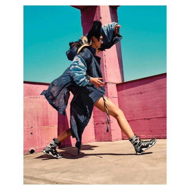 Fashion shot of a woman in avant-garde clothing with gray sneakers posing in front of pink concrete wall under bright blue sky.