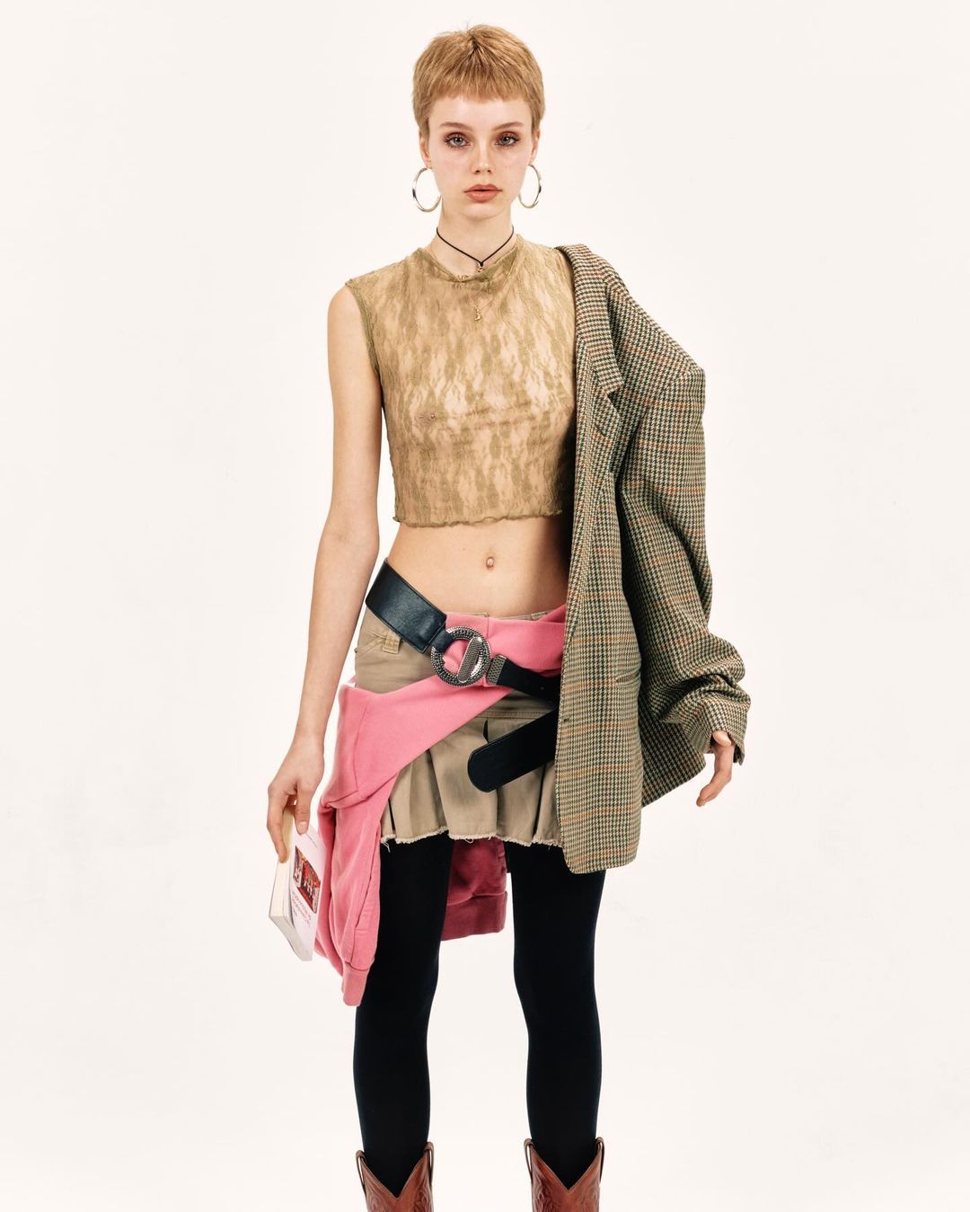 Fashionable young woman poses in a beige crop top and mini skirt with black tights and cowboy boots against a white backdrop.