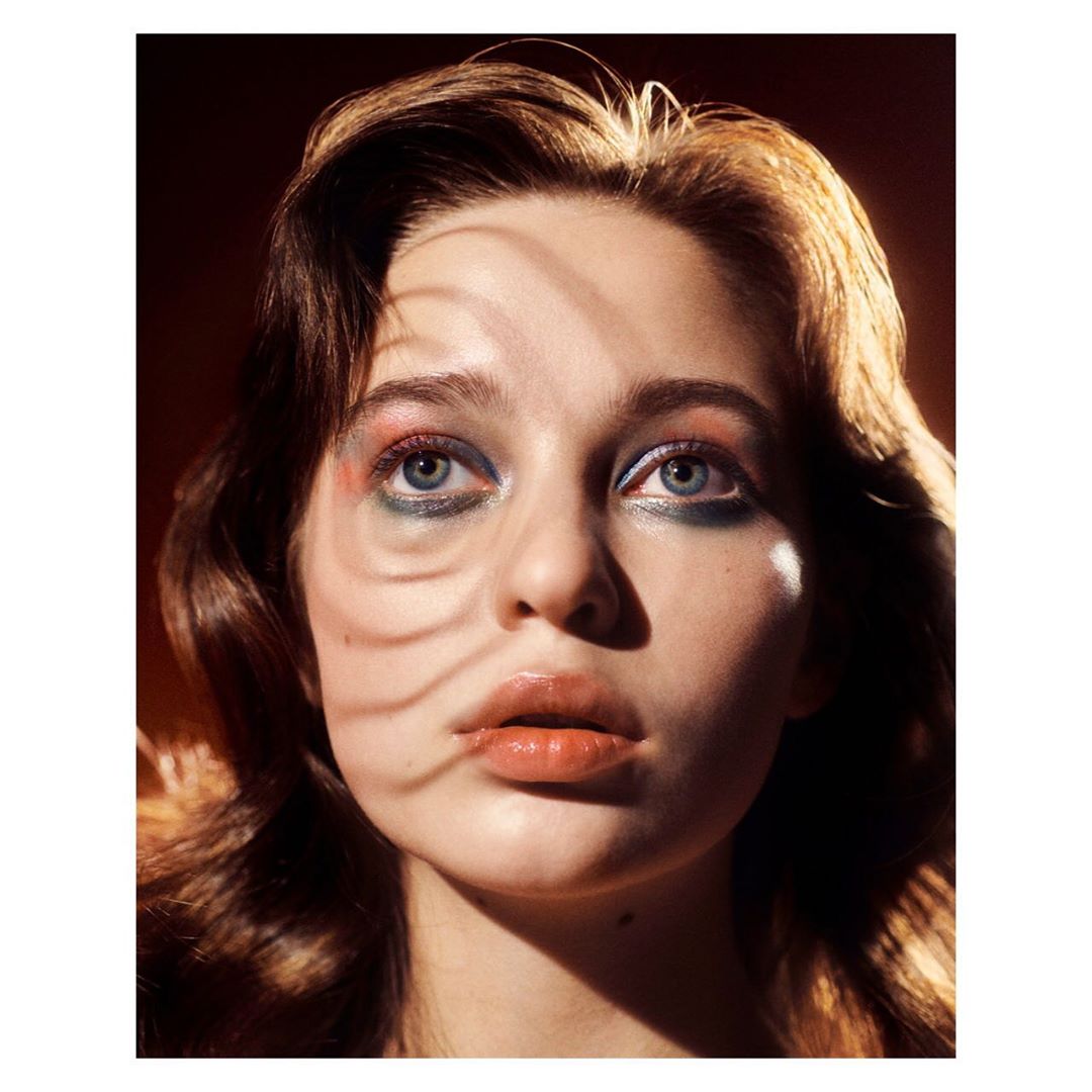 Headshot of a young woman with dramatic makeup and wavy brown hair, posing under a leaf shadow against a warm, dark background.