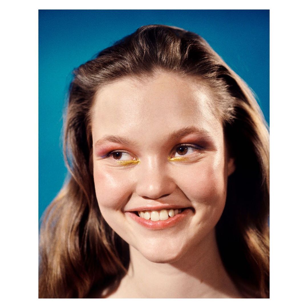 Smiling young woman with colorful makeup poses against a vibrant blue backdrop in a beauty or fashion editorial style shot.
