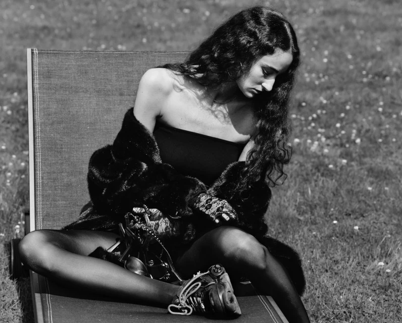 Black and white image of a woman with long curly hair seated outdoors wearing a dark tube top, fur coat, and hosiery with heels.