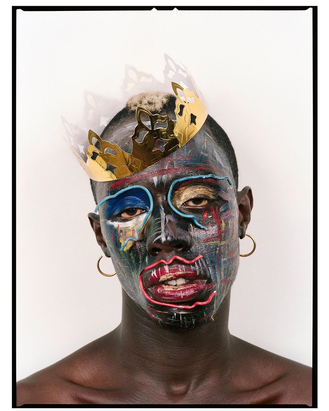 Portrait of a person with dark skin, painted face, gold crown, gold hoop earrings against a solid white backdrop in a photo studio.