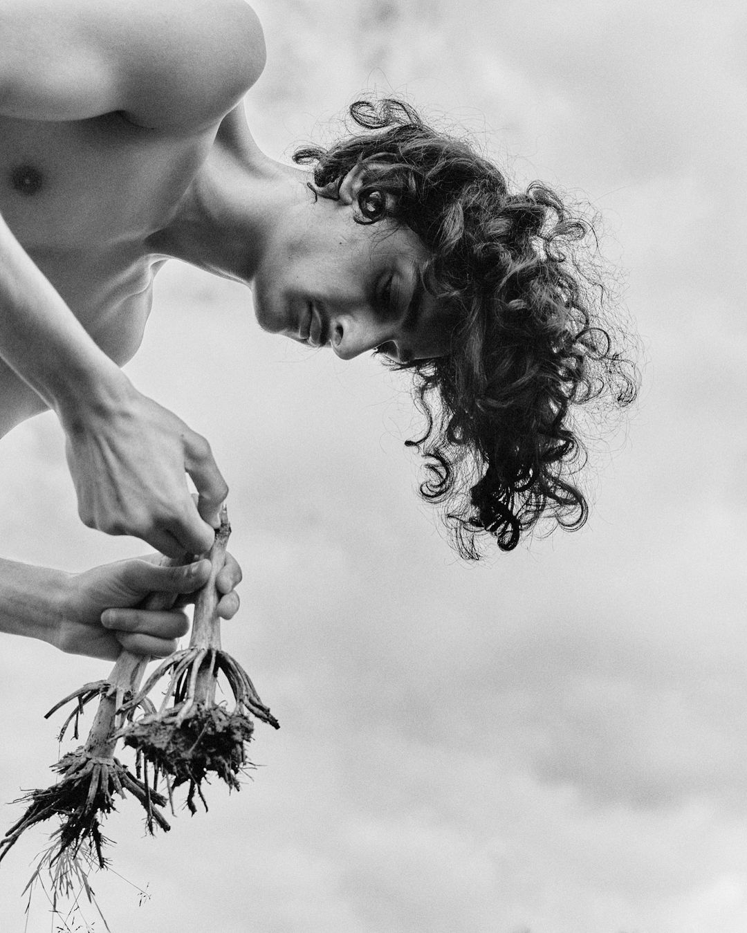 Monochrome portrait of shirtless man with curly hair holding uprooted plant against sky background, artistic and contemplative.