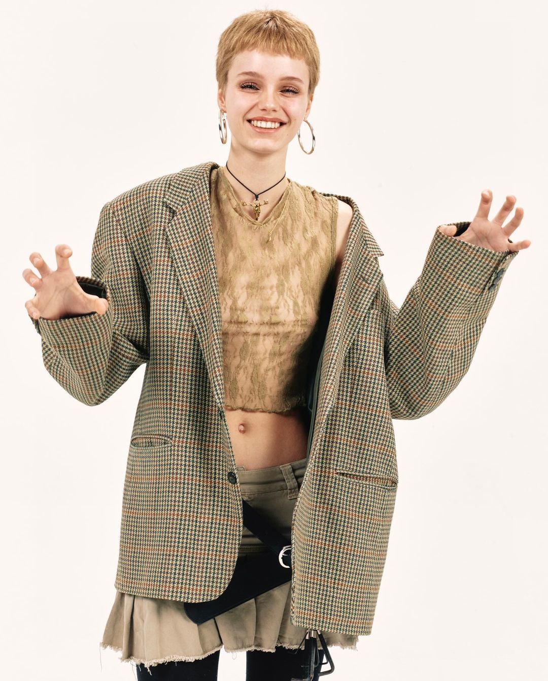 A cheerful woman with short blonde hair poses in a tan crop top, pleated mini skirt, and oversized plaid blazer against a white backdrop.