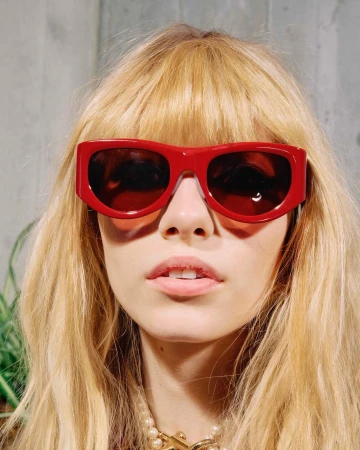Close-up portrait of a blonde woman wearing bold red sunglasses and a pearl necklace against a textured grey background.