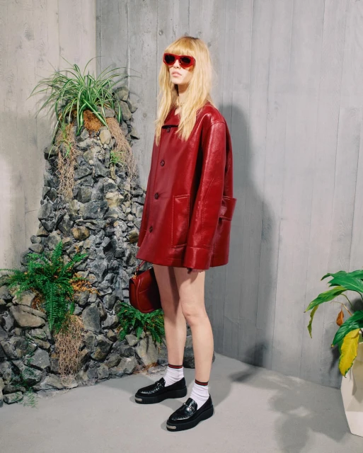 A woman poses in a red jacket with a handbag, black shoes, and sunglasses in an indoor setting with stone and plants.