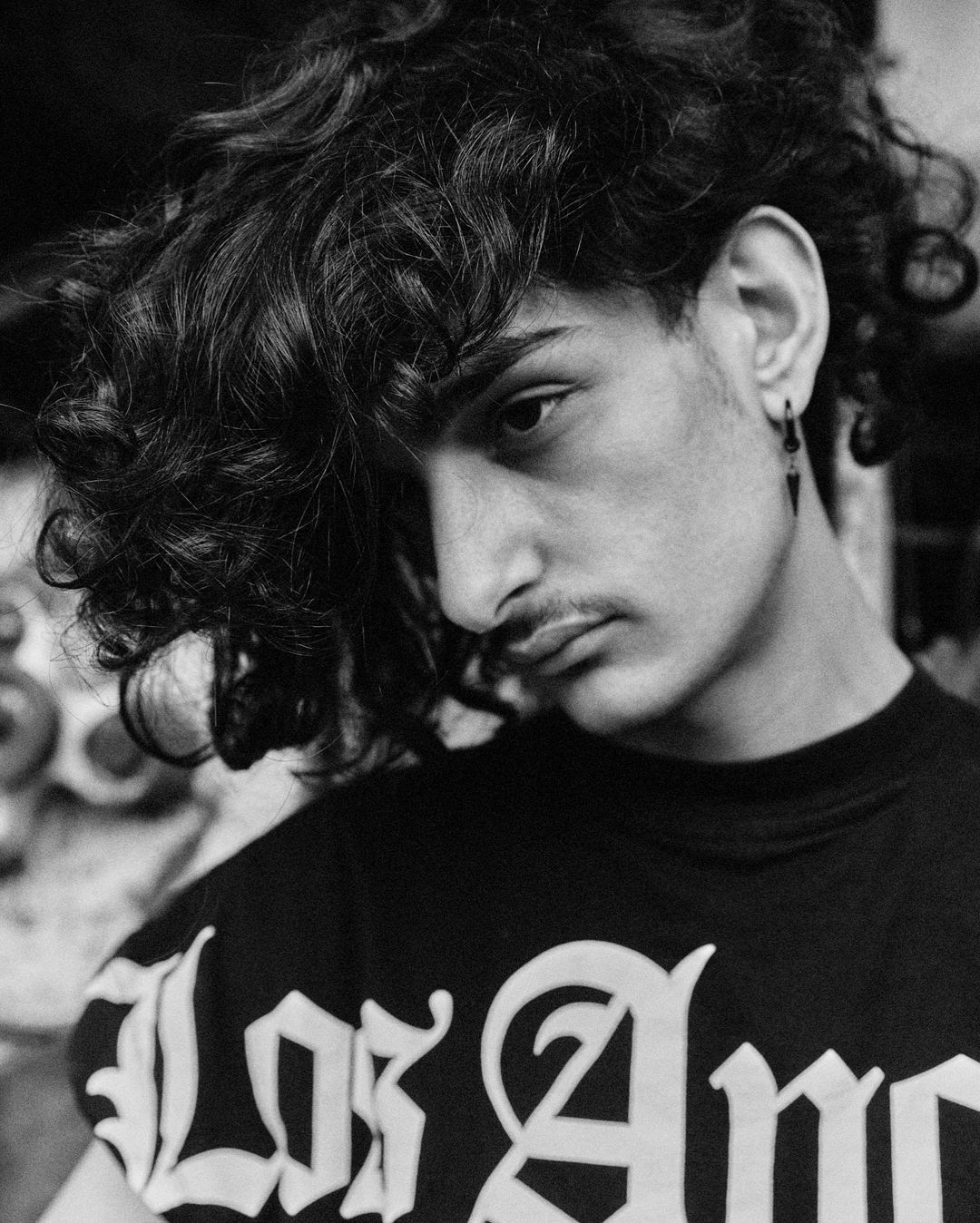 Black and white portrait of a young adult man with dark curly hair wearing a 'Los Angeles' t-shirt indoors, eye-level angle.