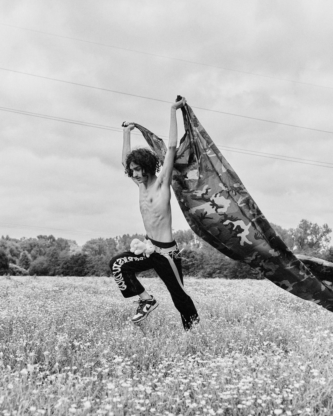 Young man jumps in a field, holding camouflage fabric. He wears dark pants with white designs and black and white sneakers in black and white.