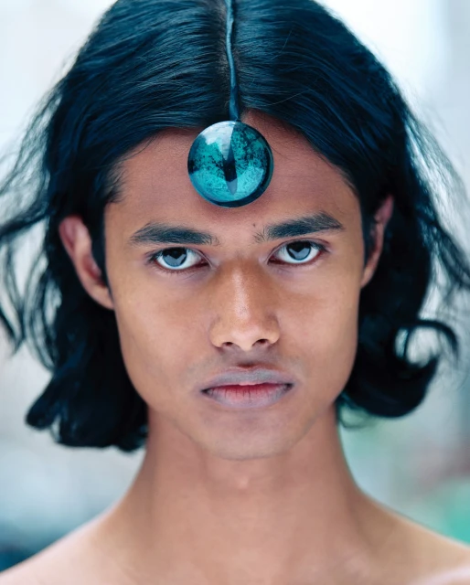 Close-up portrait of an adult male with long dark hair and a round turquoise ornament on his forehead, gazing directly at the viewer.