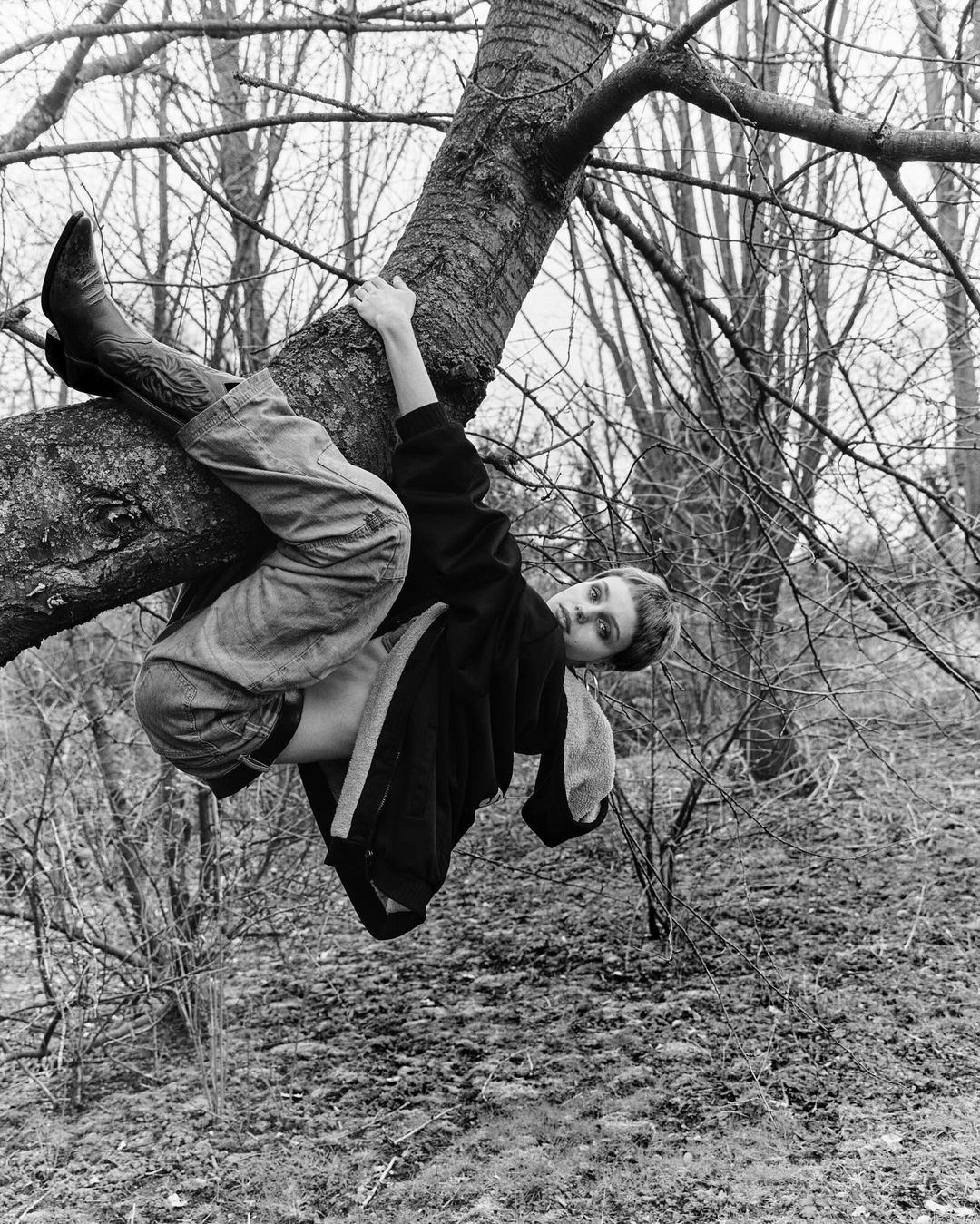 A young adult woman in cowboy boots reclines on a tree branch in a black-and-white outdoor fashion portrait within a sparse wooded area.