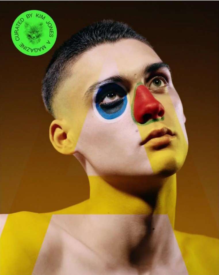 Portrait of a person with a unique face painting in red, yellow, and blue, against a warm, dark yellow background in studio lighting.