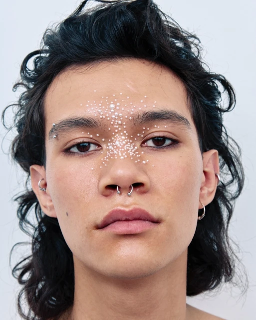 Close-up portrait of a young adult male with dark hair and decorative pearl-like makeup on his face against a plain white background.
