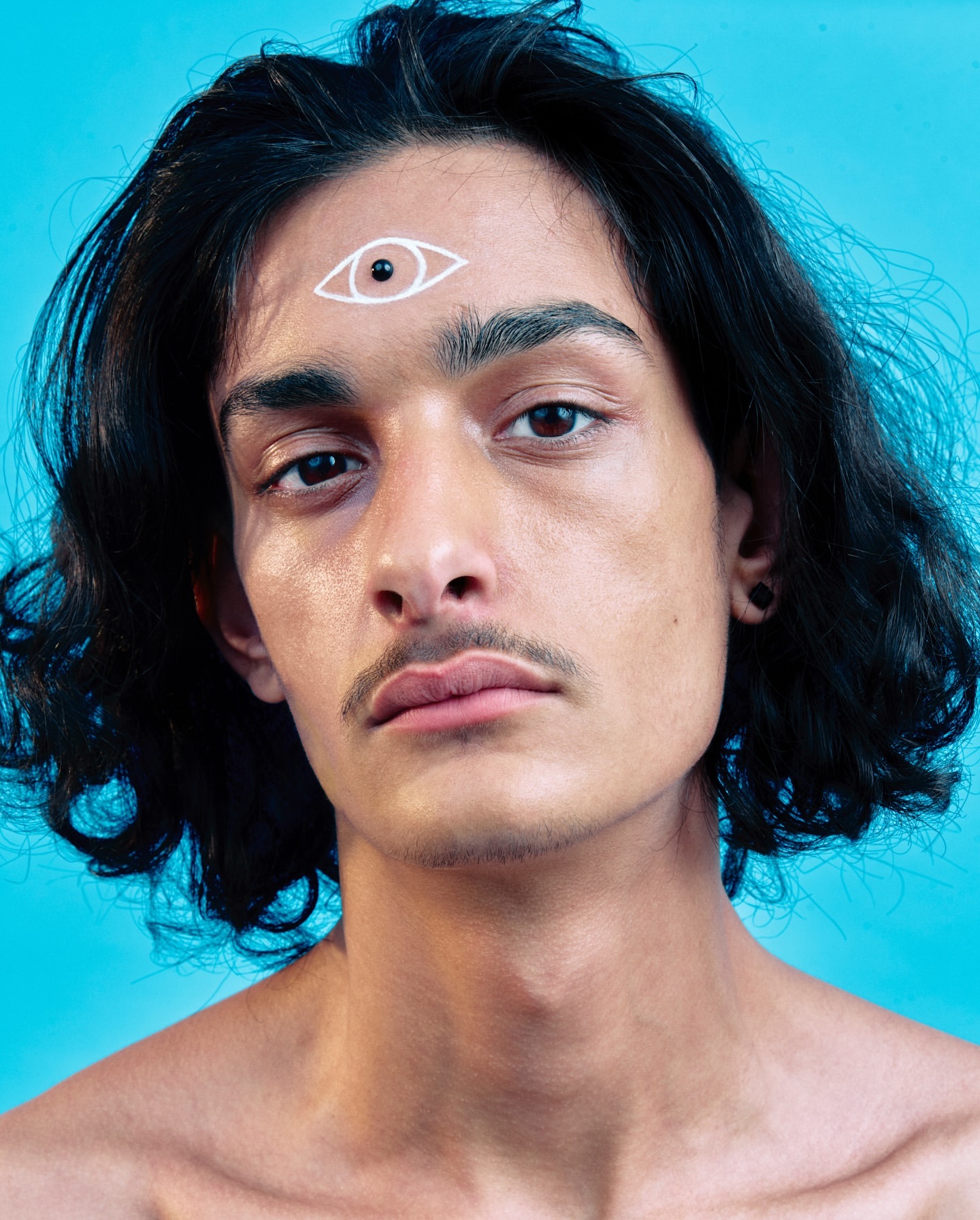 Close-up portrait of a young adult with long dark hair, mustache and a drawn third eye on their forehead against a blue background.