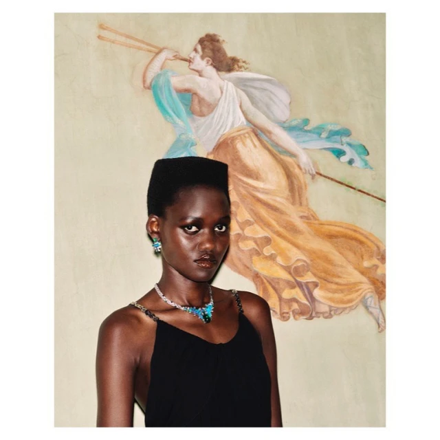Portrait of an African woman wearing a black dress, earrings, and necklace, with a painting of a woman with a staff in the background.