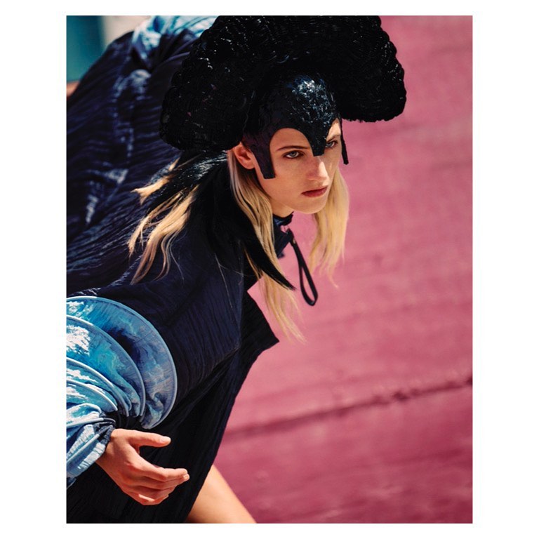 Fashion shot of a model wearing a black headdress and blue top, posing against a pink wall in an outdoor setting.