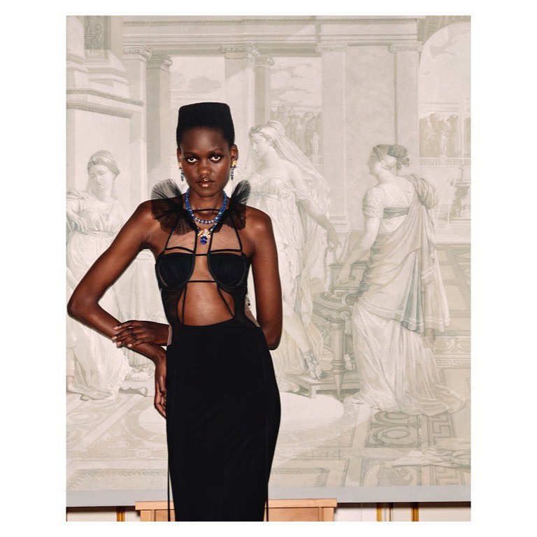 Elegant woman in a black halter dress with cutouts and tulle shoulder ruffles posing in front of a classical mural backdrop.