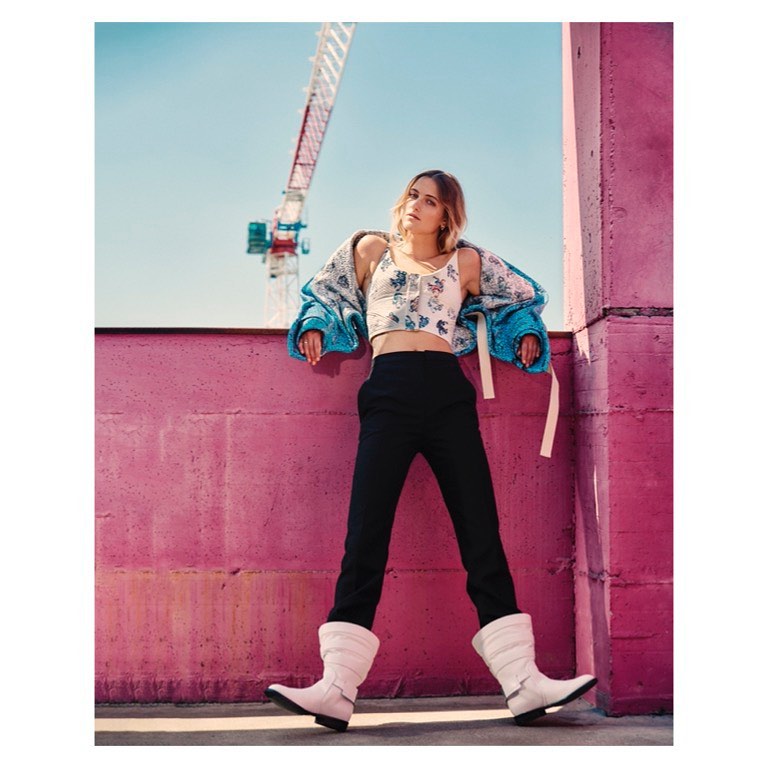 A woman in a crop top, jacket, pants and boots poses against a pink wall, with a crane visible in the background under a blue sky.