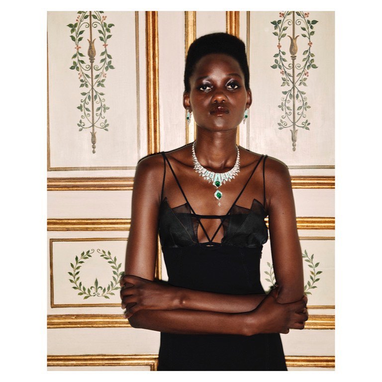 Elegant black woman in a black dress and gemstone jewelry stands against an ornate wall. Close-up studio portrait. Beauty and fashion photography.