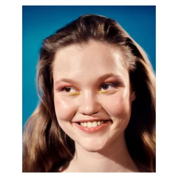 Close-up portrait of a smiling young woman with warm brown hair and colourful eye makeup against a bright blue background