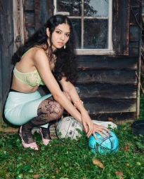Woman in light green bralette and blue skirt crouching by a wooden shed in a grassy yard, with soccer balls