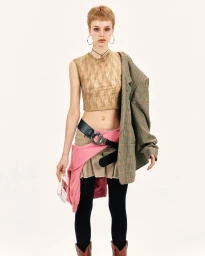 Model with short hair wearing a sheer patterned top, khaki skirt, black tights, and a houndstooth blazer draped over one shoulder.
