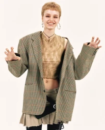 Smiling model wearing an oversized plaid blazer, sheer top, and hoop earrings against a white studio background.