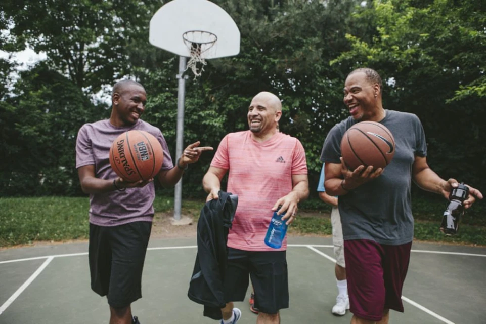 Three adult men are walking on an outdoor basketball court, smiling and holding basketballs, trees and a basketball hoop in background.